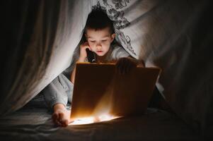 bambino lettura libro nel letto. bambini leggere a notte. poco ragazzo con Fata racconto libri nel Camera da letto . formazione scolastica per giovane bambini. di andare a dormire storia nel il sera. carino ragazzo sotto coperta nel buio camera con lampada foto