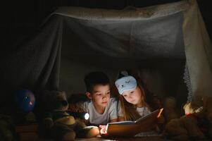 lettura e famiglia Giochi nel figli di tenda. ragazzo e ragazza con libro e torcia elettrica prima andando per letto foto