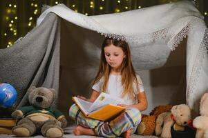 contento bambino ragazza ridendo e lettura libro nel buio nel un' tenda a casa foto