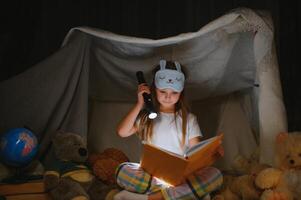 contento bambino ragazza ridendo e lettura libro nel buio nel un' tenda a casa foto