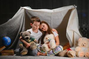 Due poco bambino giocare a casa nel il sera per costruire un' campeggio tenda per leggere libri con un' torcia elettrica e dormire dentro. foto