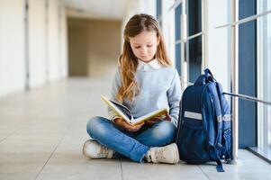 poco bella ragazza seduta su il pavimento di il scuola corridoio e lettura un' libro. il concetto di scolarizzazione. foto