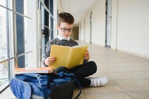 scolaro si siede su il pavimento di un' scuola corridoio e legge un' libro foto