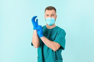 protezione contro coronavirus. metà foto di maturo maschio medico nel medico uniforme mettendo su protettivo sterile blu guanti mentre in piedi contro blu sfondo. assistenza sanitaria. coronavirus, covid19.