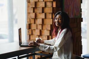 testa tiro ritratto di contento sorridente africano americano donna seduta a tavolo nel bar, guardare a telecamera, eccitato femmina in posa, Lavorando a computer, fare compiti a casa, preparazione rapporto nel caffè Casa foto