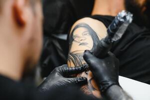 ritagliata vicino su di un' barbuto tatuaggio artista Lavorando a il suo studio tatuaggio manica su il braccio di il suo maschio cliente. uomo ottenere tatuato di professionale tatuatore foto