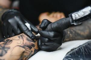 ritagliata vicino su di un' barbuto tatuaggio artista Lavorando a il suo studio tatuaggio manica su il braccio di il suo maschio cliente. uomo ottenere tatuato di professionale tatuatore foto