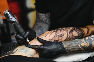 ritagliata vicino su di un' barbuto tatuaggio artista Lavorando a il suo studio tatuaggio manica su il braccio di il suo maschio cliente. uomo ottenere tatuato di professionale tatuatore foto