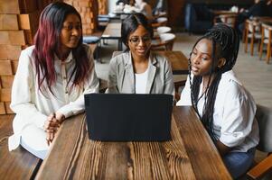 tre africano americano ragazze seduta a il tavolo nel bar studiando su per test o fabbricazione compiti a casa insieme, essi siamo utilizzando il computer portatile e digitale tavoletta. foto