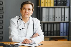 ritratto tiro di mezzo anziano femmina medico seduta a scrivania e Lavorando nel ufficio. foto