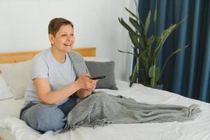 di mezza età donna seduta su il divano e svolta su il tv con un' a distanza controllo. foto