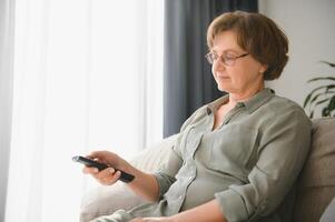 tecnologia, televisione, età e persone concetto - contento anziano donna Guardando tv e mutevole canale con a distanza controllo a casa foto