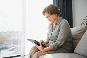 anziano donna utilizzando digitale tavoletta nel vivente camera a casa foto