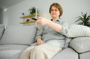 tecnologia, televisione, età e persone concetto - contento anziano donna Guardando tv e mutevole canale con a distanza controllo a casa foto