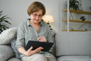 anziano donna utilizzando digitale tavoletta nel vivente camera a casa foto