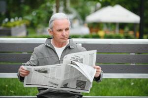 bello nonno si siede su un' panchina nel il parco e legge un' giornale. anziano dai capelli grigi uomo. foto