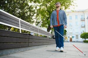 giovane cieco uomo con bianca canna a piedi attraverso il strada nel città foto