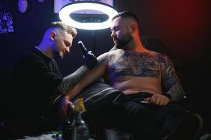 barbuto tatuaggio artista Lavorando a il suo studio tatuaggio manica su il braccio di il suo maschio cliente. uomo ottenere tatuato di professionale tatuatore foto