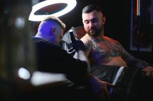 barbuto tatuaggio artista Lavorando a il suo studio tatuaggio manica su il braccio di il suo maschio cliente. uomo ottenere tatuato di professionale tatuatore foto