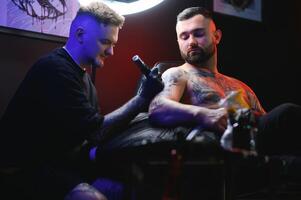 barbuto tatuaggio artista Lavorando a il suo studio tatuaggio manica su il braccio di il suo maschio cliente. uomo ottenere tatuato di professionale tatuatore foto