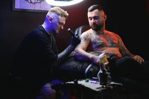 barbuto tatuaggio artista Lavorando a il suo studio tatuaggio manica su il braccio di il suo maschio cliente. uomo ottenere tatuato di professionale tatuatore foto