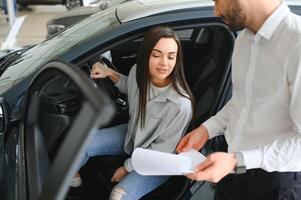 sorridente auto venditore in piedi nel auto salone con cliente e mostrando in giro macchine su vendita foto