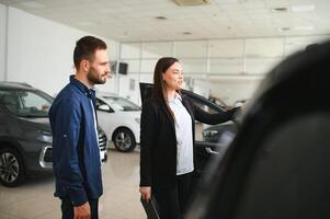 amichevole, sorridente femmina venditore mostrando marca nuovo auto per un' cliente mentre in piedi nel auto salone foto