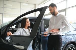 soddisfatto contento caucasico femmina cliente cliente donna seduta a il ruota di nuovo auto mentre maschio negozio assistente porzione sua scegliere esso foto