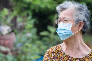 un anziano donna indossare un' chirurgico maschera e guardare lontano mentre in piedi nel un' giardino. maschera per proteggere virus, coronavirus, covid19, e di più. concetto di anziano persone e assistenza sanitaria foto