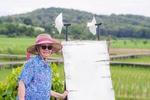 anziano donna indossare cappello e occhiali da sole in piedi accanto un' vuoto bianca di legno cartello con un' sfondo di riso i campi e montagne. spazio per testo. concetto di anziano persone e rilassamento foto