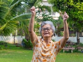 un' contento anziano donna mostrando stringendo pugni eccitato per successo con braccia sollevato festeggiare vittoria sorridente mentre in piedi nel il giardino. concetto di anziano persone e assistenza sanitaria foto