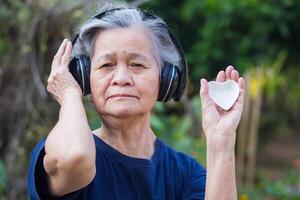 anziano donna con corto grigio capelli, indossare senza fili cuffia, Tenere un' a forma di cuore bianca ceramica, e guardare a il telecamera mentre in piedi nel un' giardino. concetto di anziano persone e rilassamento foto