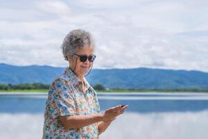 ritratto di anziano donna indossare occhiali da sole Tenere e guardare a un' smartphone mentre in piedi a il lato di il lago con montagna sfondo. concetto di anziano persone e tecnologia foto