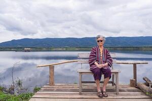 ritratto di un anziano asiatico donna indossare occhiali da sole mentre seduta su un' di legno sedia lato il lago. concetto di anziano persone e rilassamento foto