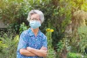 anziano donna indossare un' viso maschera e braccia attraversato mentre in piedi nel un' giardino. maschera per proteggere covid19, virus, coronavirus, polline cereali, eccetera. concetto di anziano persone e assistenza sanitaria foto