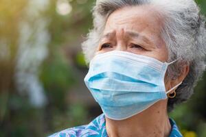 anziano donna indossare un' viso maschera e guardare su mentre in piedi nel un' giardino. maschera per proteggere covid19, virus, coronavirus, polline cereali, eccetera. concetto di anziano persone e assistenza sanitaria foto