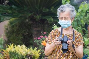 anziano donna indossare un' viso maschera e tiro un' foto di il digitale telecamera mentre in piedi nel un' giardino. maschera per proteggere virus, coronavirus. nuovo normale. concetto di anziano persone e fotografia