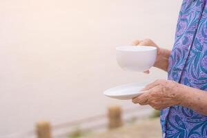 avvicinamento di vecchio donna mani hold un' bianca caffè tazza mentre in piedi a il lato il fiume. mattina caffè. spazio di testo. concetto di anziano persone e rilassamento foto