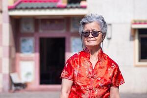 asiatico anziano donna indossare un' tradizionale cheongsam qipao vestito e occhiali da sole, guardare a il telecamera mentre in piedi a Cinese tempio. concetto di anziano persone e Cinese nuovo anno foto