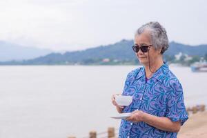 un anziano donna con corto grigio capelli Tenere un' bianca caffè tazza mentre in piedi lato il fiume con natura sfondo. mattina caffè. spazio di testo concetto di anziano persone e rilassamento foto