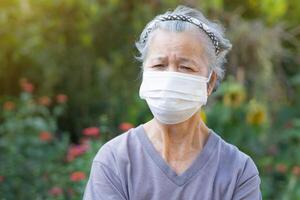 un anziano donna indossare un' chirurgico maschera mentre in piedi nel un' giardino. maschera per proteggere virus, coronavirus, covid19, batteri, polline cereali, e di più. concetto di anziano persone e assistenza sanitaria foto