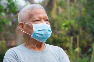un' ritratto di un anziano uomo indossare un' viso maschera guardare lontano mentre in piedi nel un' giardino. maschera per proteggere virus, covid19, coronavirus, batteri, e di più. concetto di anziano persone e assistenza sanitaria foto