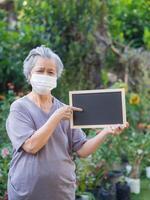 ritratto di anziano donna in piedi Tenere un' nero etichetta e indossare viso maschera per salutare perché avere aria inquinamento pm 2.5. maschera per proteggere virus, batteri, polline grani. assistenza sanitaria concetto foto