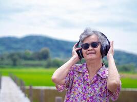 anziano donna indossare senza fili cuffie ascoltando per musica mentre in piedi lato riso i campi con montagne sfondo. spazio per testo. concetto di anziano persone e rilassamento foto