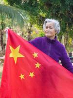 ritratto di anziano donna con corto bianca capelli Tenere Cinese bandiera, in piedi nel giardino, in piedi nel sua giardino. foto