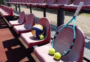 un' tennis racchetta, palle e un' pallavolo su un' panchina su il gli sport tribuna foto