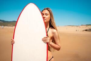 attraente giovane donna in posa con tavola da surf su sabbioso spiaggia foto