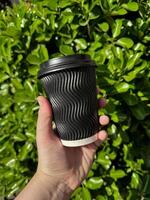 mano Tenere nero ondulato cartone caffè tazza con coperchio contro sfondo di lussureggiante verde frondoso cespuglio. urbano eco amichevole stile di vita. vuoto spazio per etichetta o etichetta. foto