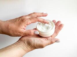 vicino su di mezzo età donna mani Tenere contenitore di crema e immersione dito su bianca sfondo. cura della pelle e idratante concetto. per assistenza sanitaria, benessere, e se stesso cura manifesto. foto