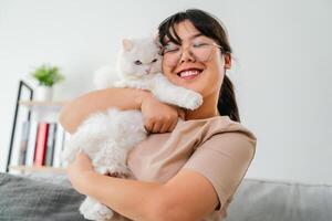 un' donna è Tenere un' bianca gatto e sorridente nel il vivente camera a casa. foto